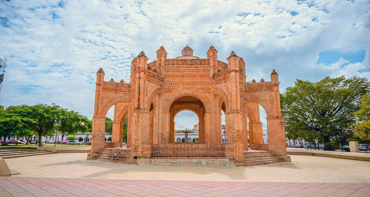 Pabellón de fuente de ladrillo de estilo morisco en una plaza abierta bajo un cielo parcialmente nublado.
