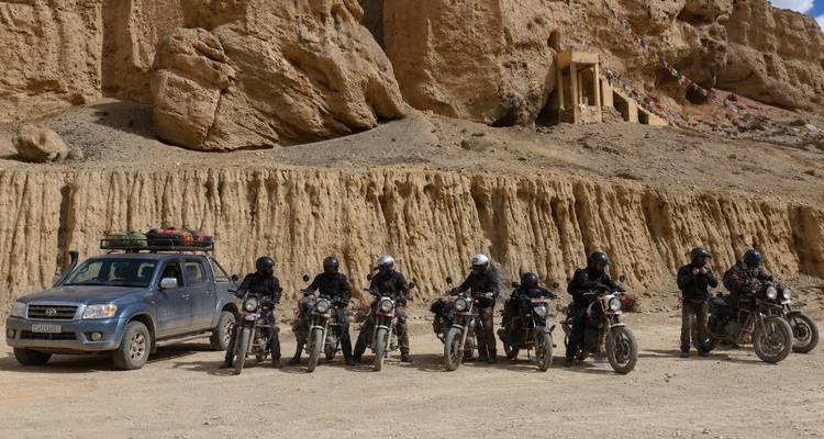 A convoy of motorcyclists and a support truck pause beneath towering, eroded cliffs in a high-desert valley.