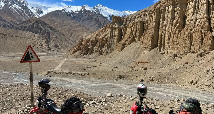 Two motorcycles parked beside a barren valley with snow-capped peaks and dramatic rock formations.