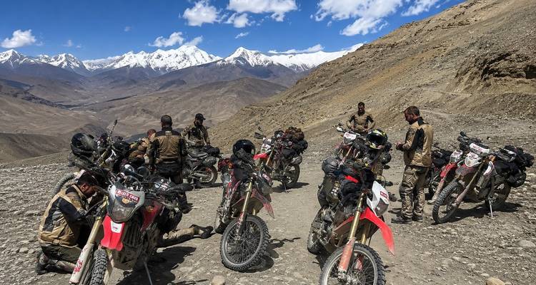 A group of off-road motorcyclists rest on a rocky mountain pass with snowy peaks beyond.