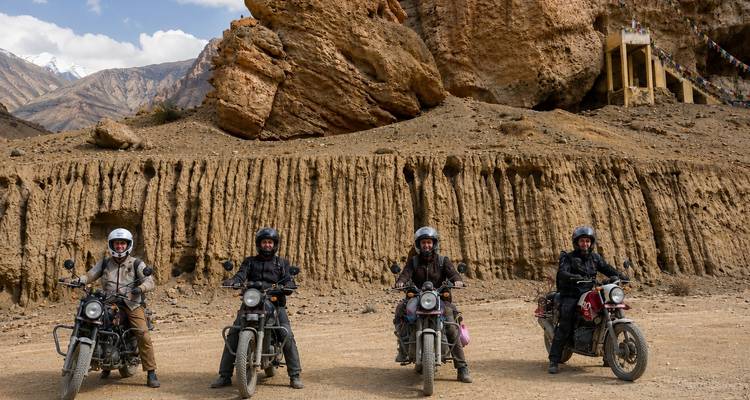 Four motorcyclists lined up with classic bikes on a dusty Himalayan plateau beneath eroded cliffs.