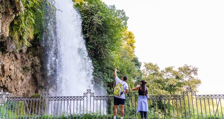 Un couple pointe vers une haute cascade qui plonge le long d'une falaise boisée
