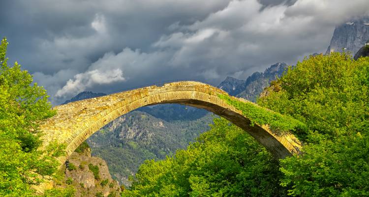 Un pont en arc de pierre solitaire drapé de verdure enjambe un ravin forestier profond sous des nuages orageux.