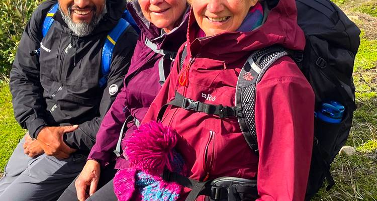 Tres senderistas con chaquetas coloridas se sientan juntos descansando en el suelo cubierto de pasto durante una caminata.