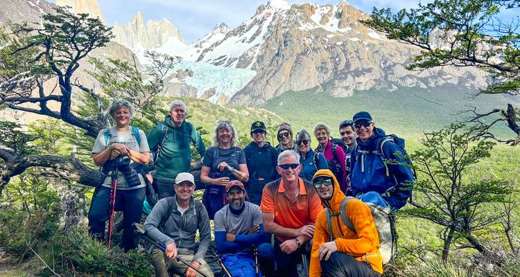 Grupo de senderismo se reúne en un claro del bosque con un glaciar colgante y picos dentados visibles más allá de los árboles.