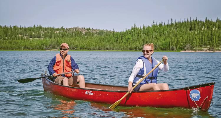 Zwei Personen paddeln in einem Kanu auf einem ruhigen See, umgeben von Bäumen.