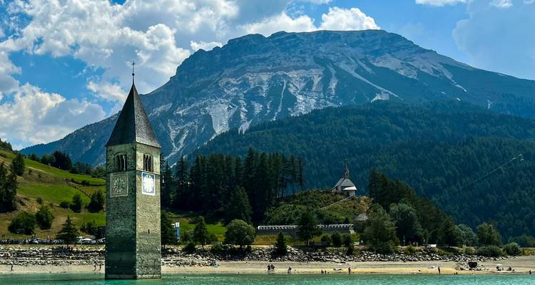 Torre de iglesia hundida en un lago con montañas al fondo.