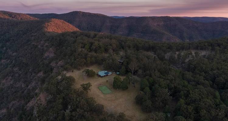 Vista aérea de una cabaña remota rodeada de montañas boscosas al atardecer.