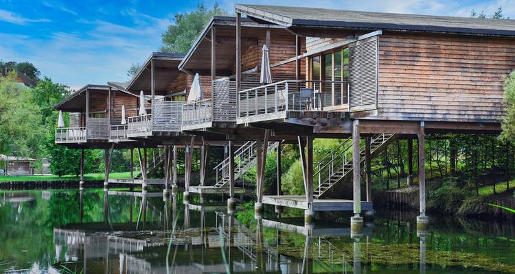 Maisons en bois sur pilotis au-dessus d'un lac calme entouré de verdure.