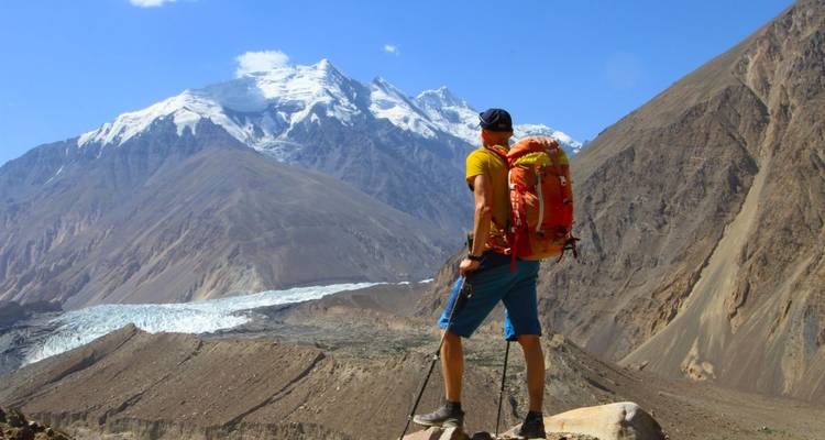 Wanderer steht mit Rucksack auf einem Felsen und blickt auf schneebedeckte Berge.