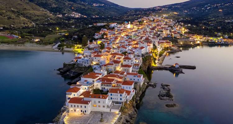 Luftaufnahme eines griechischen Küstendorfs bei Nacht mit Lichtern, die sich auf dem Meer spiegeln.