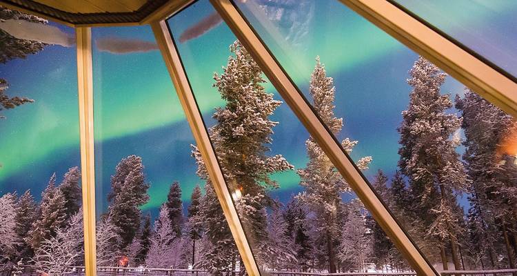 Auroras boreales vistas a través de una cabaña con techo de cristal con pinos cubiertos de nieve creando una escena mágica de invierno.