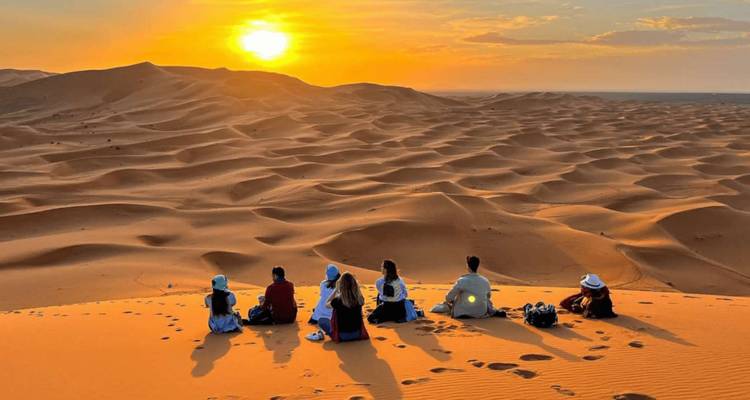 A line of travellers sitting on a dune watching the sun set over vast rolling Sahara sand dunes.