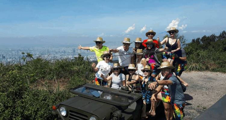 Group of tourists posing with a military jeep overlooking a city and mountains in the distance.