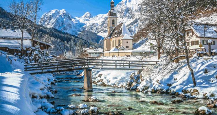 Un village avec un pont en bois au-dessus d'une rivière, entouré de montagnes enneigées.