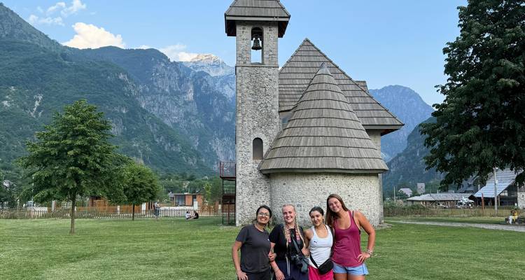 Personas posando frente a una pequeña iglesia de piedra con montañas.
