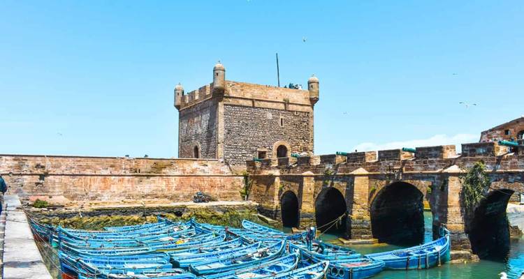 Port d'Essaouira avec des bateaux bleus amarrés près d'une forteresse en pierre.