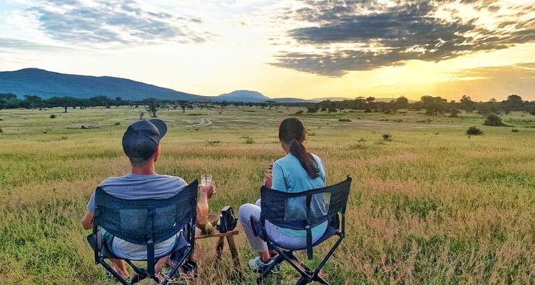 Zwei Personen sitzen in Stühlen mit Blick auf eine Savannenlandschaft bei Sonnenuntergang.