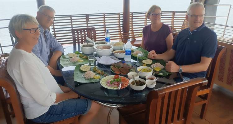 Reizigers genieten van een lunch op een bananenblad aan een ronde glazen tafel met uitzicht op water.