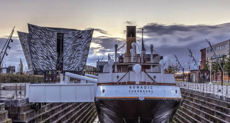 Het Nomadic schip aan de kade met moderne architectuur op de achtergrond.
