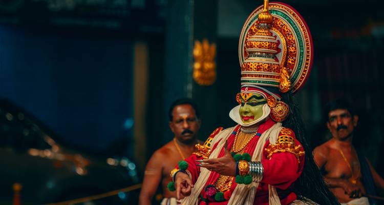 Danseur Kathakali vibrant en costume complet se produisant sur scène, observé par des musiciens.