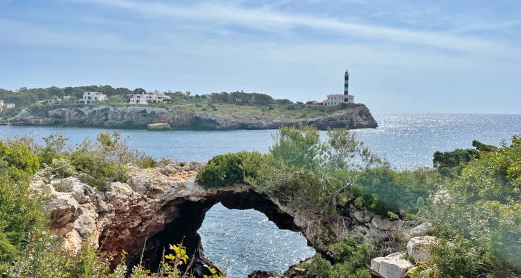 Un arco marino natural en primer plano enmarca un faro distante en un islote rocoso bajo un mar resplandeciente.