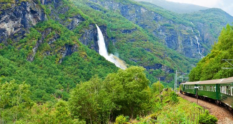 Train with green cars passing through lush valley with waterfalls.