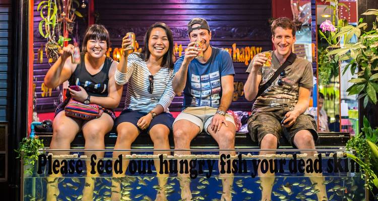 Vier glimlachende reizigers zitten met hun voeten in een visfspa tank en houden drankjes vast onder kleurrijke nachtleven verlichting.