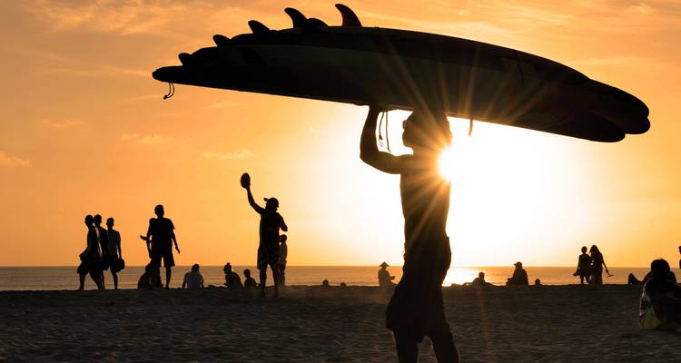 Silueta de personas en una playa durante el atardecer, con una persona cargando una tabla de surf.