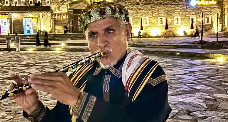 Man in traditional attire playing a flute in front of a cultural backdrop.