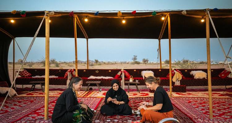Three women sitting inside a tent placed in a desert landscape.