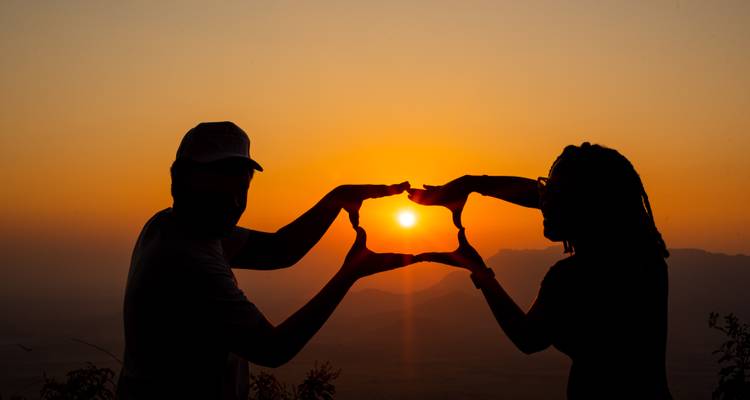 Silhouette von zwei Personen, die die untergehende Sonne mit ihren Händen einrahmen.