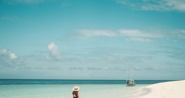 Une femme debout sur une plage immaculée avec un boutre à l'horizon.