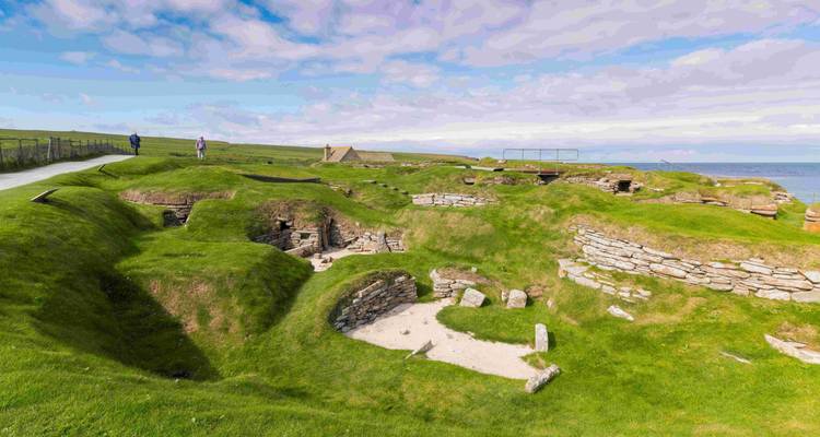 Ancient stone structures on grassy mounds with ocean in the background.