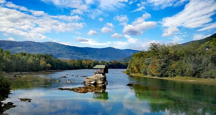 Une petite maison sur un rocher dans une rivière avec des collines environnantes et un ciel bleu.