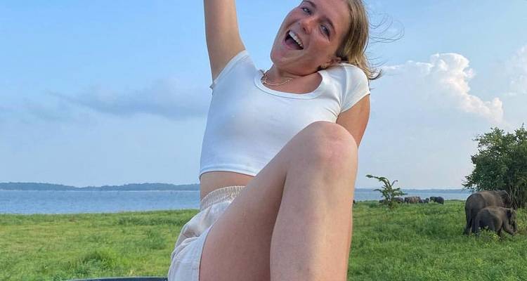 Cheerful person sitting on a grassy field near a lake with elephants in the background.