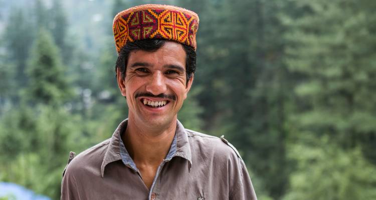 Hombre sonriente usando un gorro Himachali colorido con un fondo de colinas de bosque de pinos