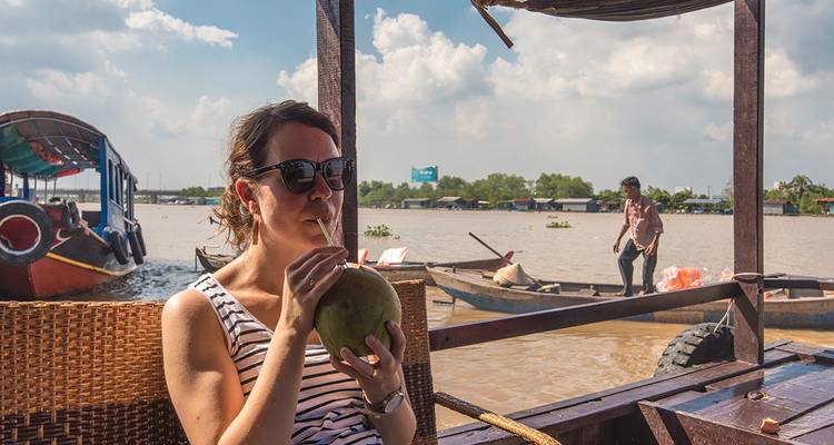Mujer bebiendo coco en un barco, con gente y barcos en un río.