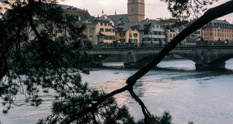 Paysage urbain avec un pont historique et une tour, vu depuis la berge d'une rivière.