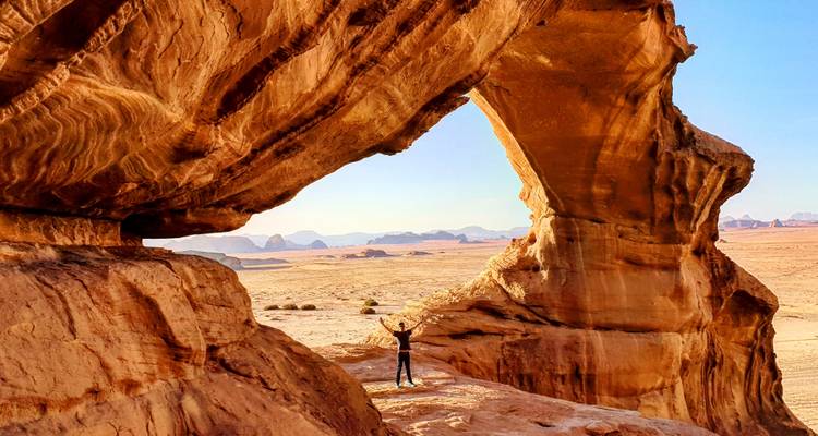 Persona parada bajo un gran arco de roca en un desierto.