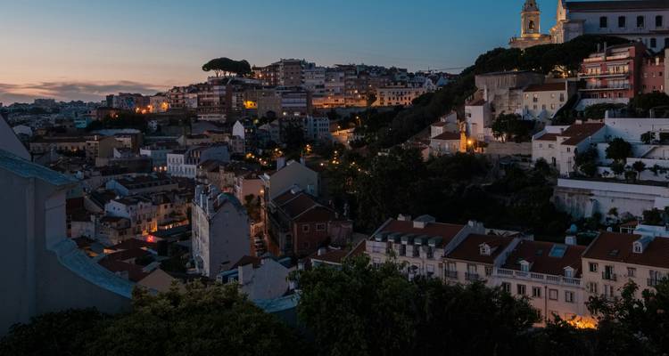 De heuvels van Lissabon gloeien bij schemering in warm licht, met dakpannen en kerktorens die boven de smalle straatjes uitsteken.