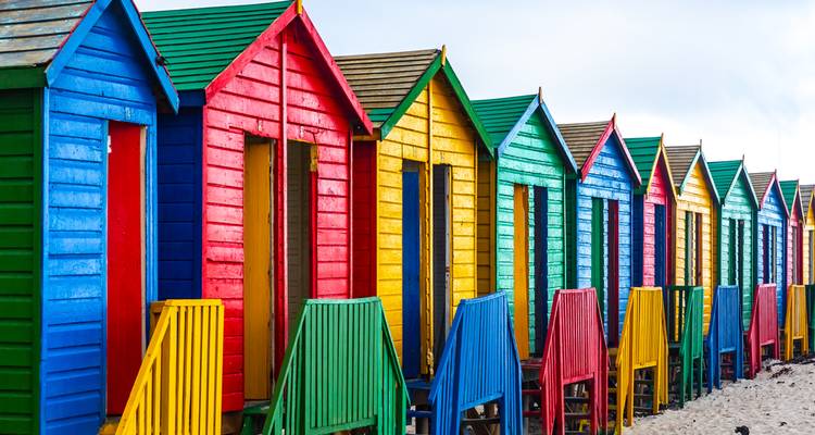 Una fila de casetas de playa de madera pintadas de colores brillantes en vivos colores del arcoíris se alinea en una orilla arenosa.