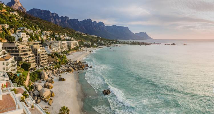 Una vista aérea panorámica de Camps Bay muestra el oleaje turquesa, la arena blanca y las montañas Twelve Apostles al atardecer.