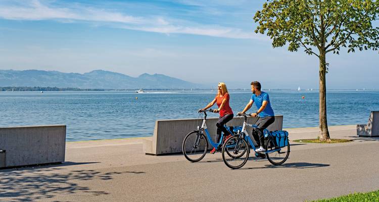 Deux personnes faisant du vélo le long d'un sentier au bord d'un lac avec des montagnes en arrière-plan.