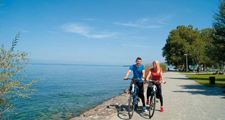 Deux personnes faisant du vélo sur un sentier au bord du lac avec un ciel bleu dégagé.