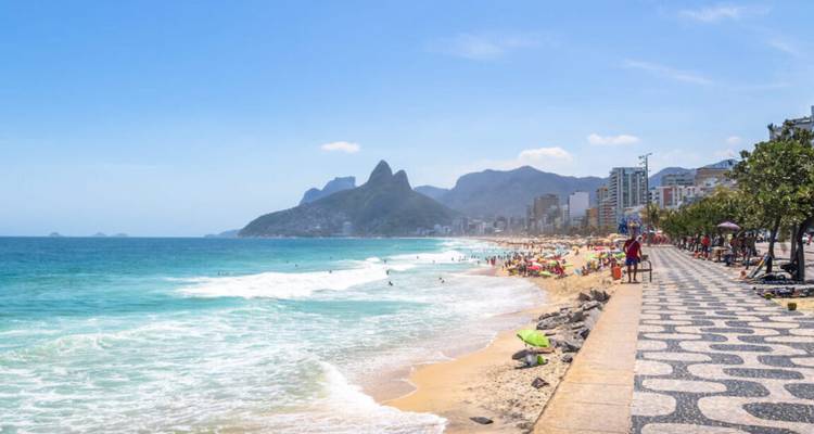 Belebter Strand von Ipanema mit gemusterter Promenade, türkisfarbenen Wellen und den Gipfeln der Dois Irmãos im Hintergrund.