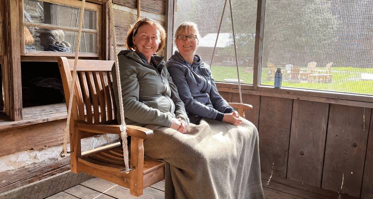 Deux femmes assises sur une balançoire de porche enveloppées dans des couvertures, souriant à la caméra.