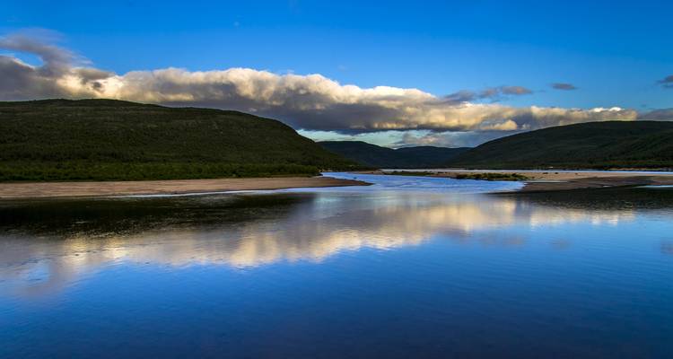 Malerische Aussicht auf einen Fluss, umgeben von Bergen und bewölktem Himmel.