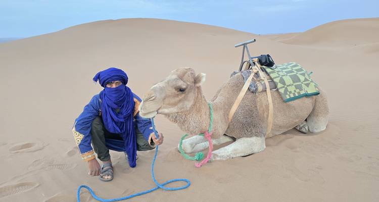Man in traditionele kledij met een kameel in de woestijn.
