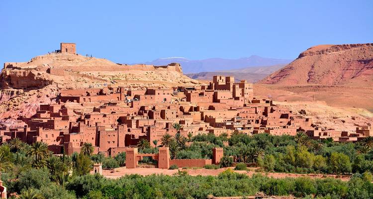Le ksar en terre iconique d'Aït Benhaddou s'élève au-dessus de palmiers luxuriants avec les montagnes de l'Atlas en arrière-plan sous un ciel bleu dégagé.